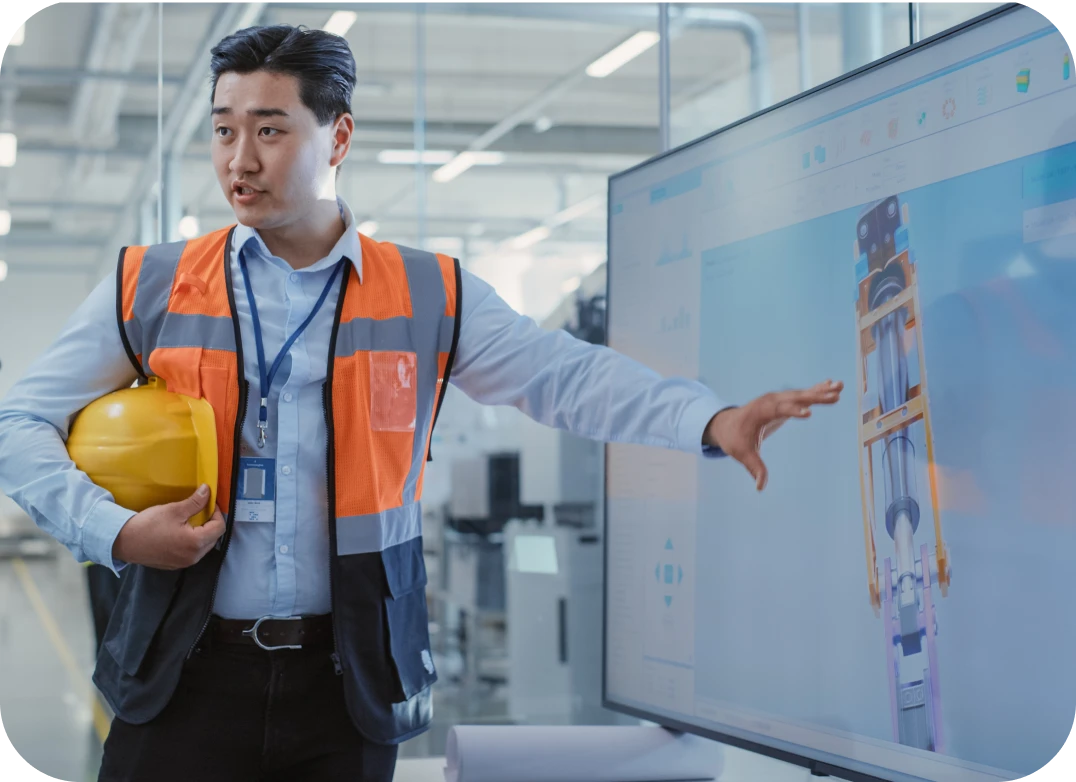 A male employee with a bright orange working vest points to a digital signage screen that is displaying a 3D model of a machine part