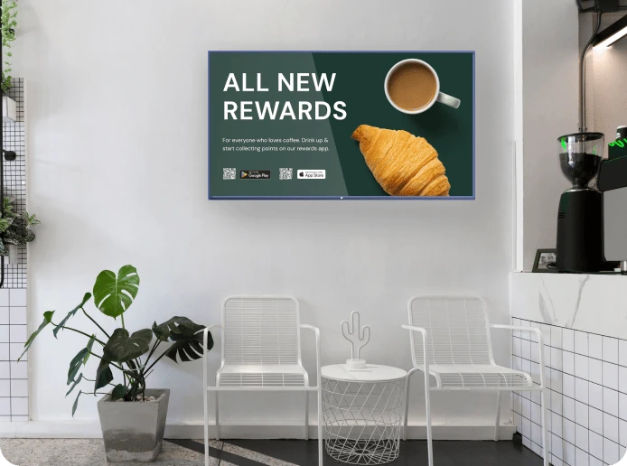 A digital signage screen on a cafe wall displays a QR code and advertisement for the cafe's rewards app