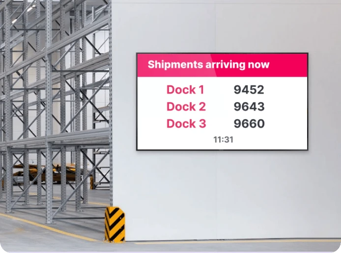 A digital signage screen mounted on an empty wall in a warehouse displays shipment arrival information in real time.